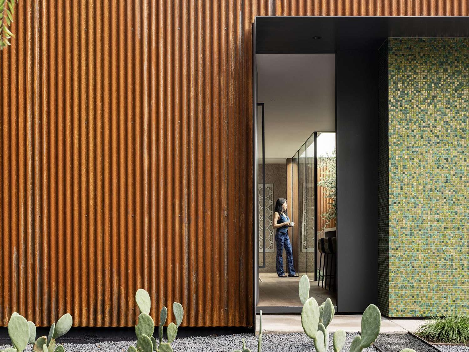 A bold mix of industrial and refined materials, this modern home pairs rusted corrugated steel with a shimmering mosaic wall for a striking first impression. The narrow window offers a glimpse into the tranquil, glass-lined interior.