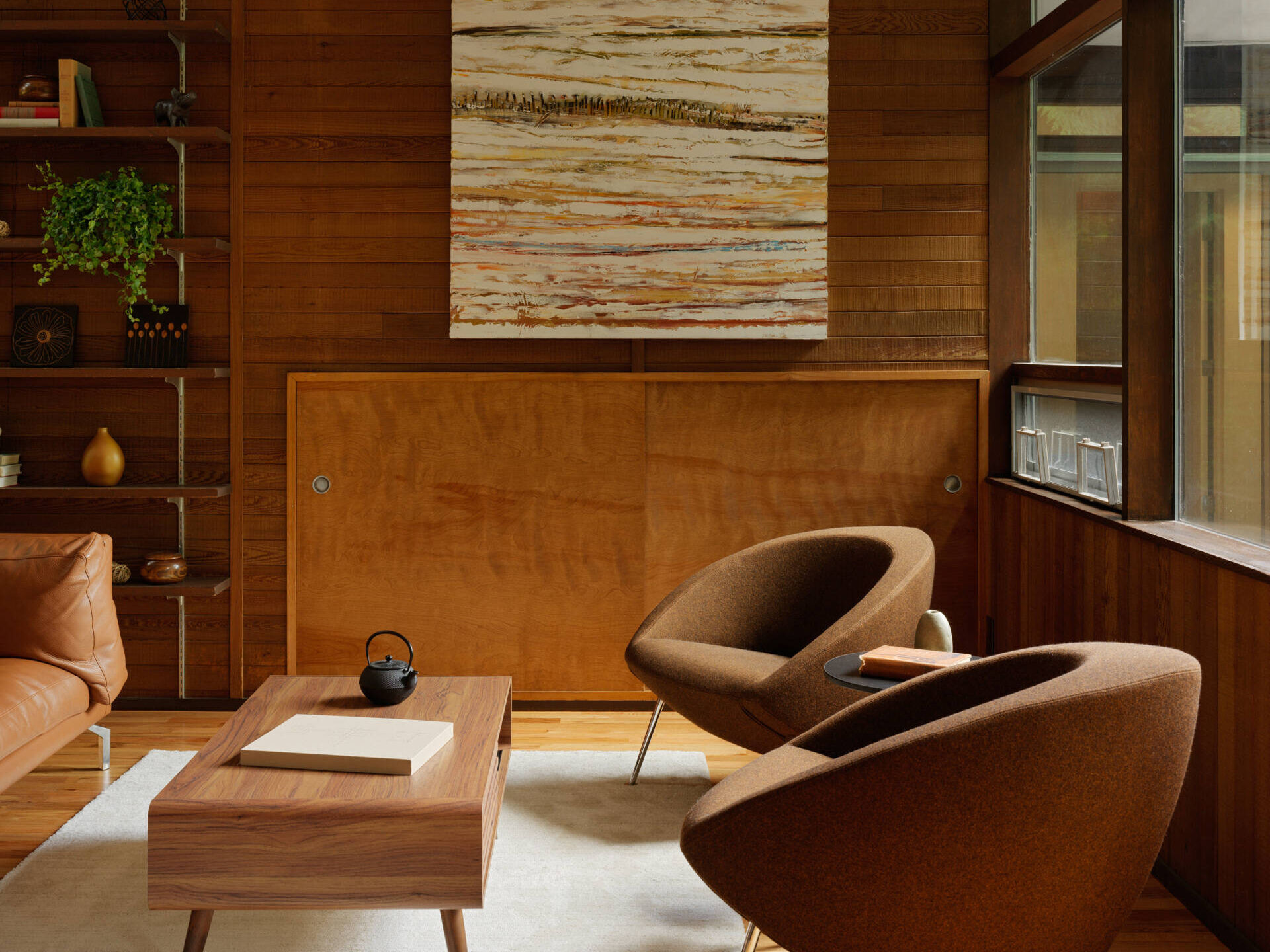 Floor-to-ceiling windows invite the surrounding trees and creek views into this mid-century modern inspired living room, while a bold steel fireplace anchors the room. Sculptural works by Alden Mason and a painting by Simeon Nikolov enrich the space with personal and artistic depth. The design encourages quiet reflection and warmth.