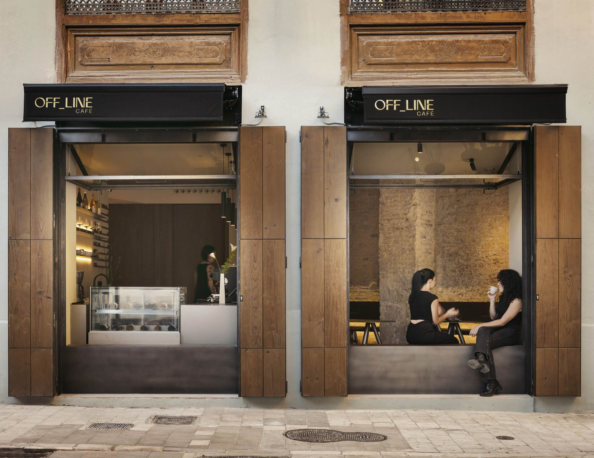 Tucked into the corner of a charming old building, this café blends urban cool with a warm neighborhood vibe. The facade makes a striking first impression, pairing natural wood shutters with crisp black signage that glows softly at dusk. Large open windows draw the eye inward, where golden light spills across exposed brick walls and minimalist black furniture. Above, carved wooden window frames and ornate wrought iron balconies hint at the building’s historic roots, adding a touch of Old World elegance to the contemporary setup below. 