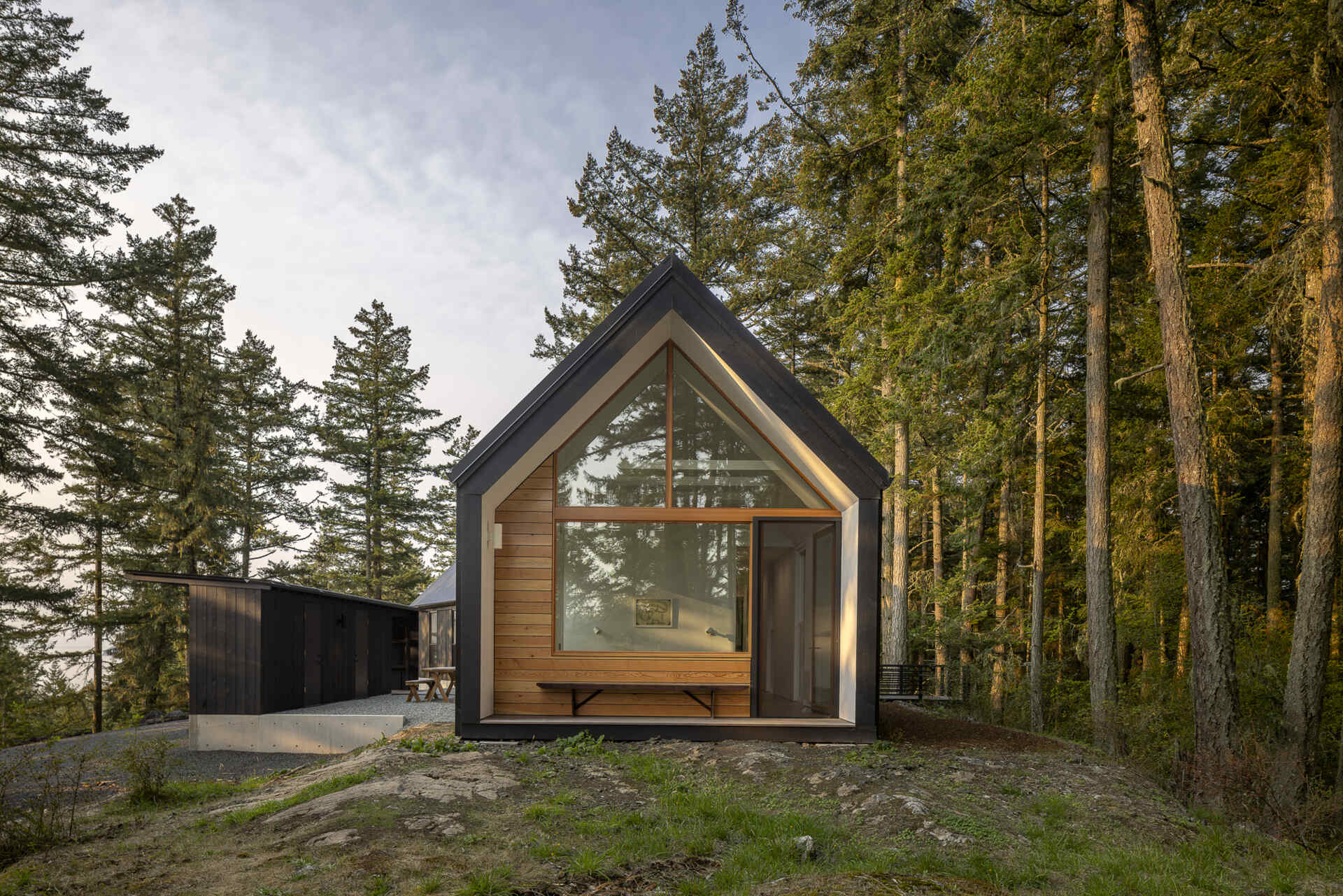 A minimalist gabled home on Orcas Island finished in dark weathered wood, set among old growth trees with layered views in every direction.
