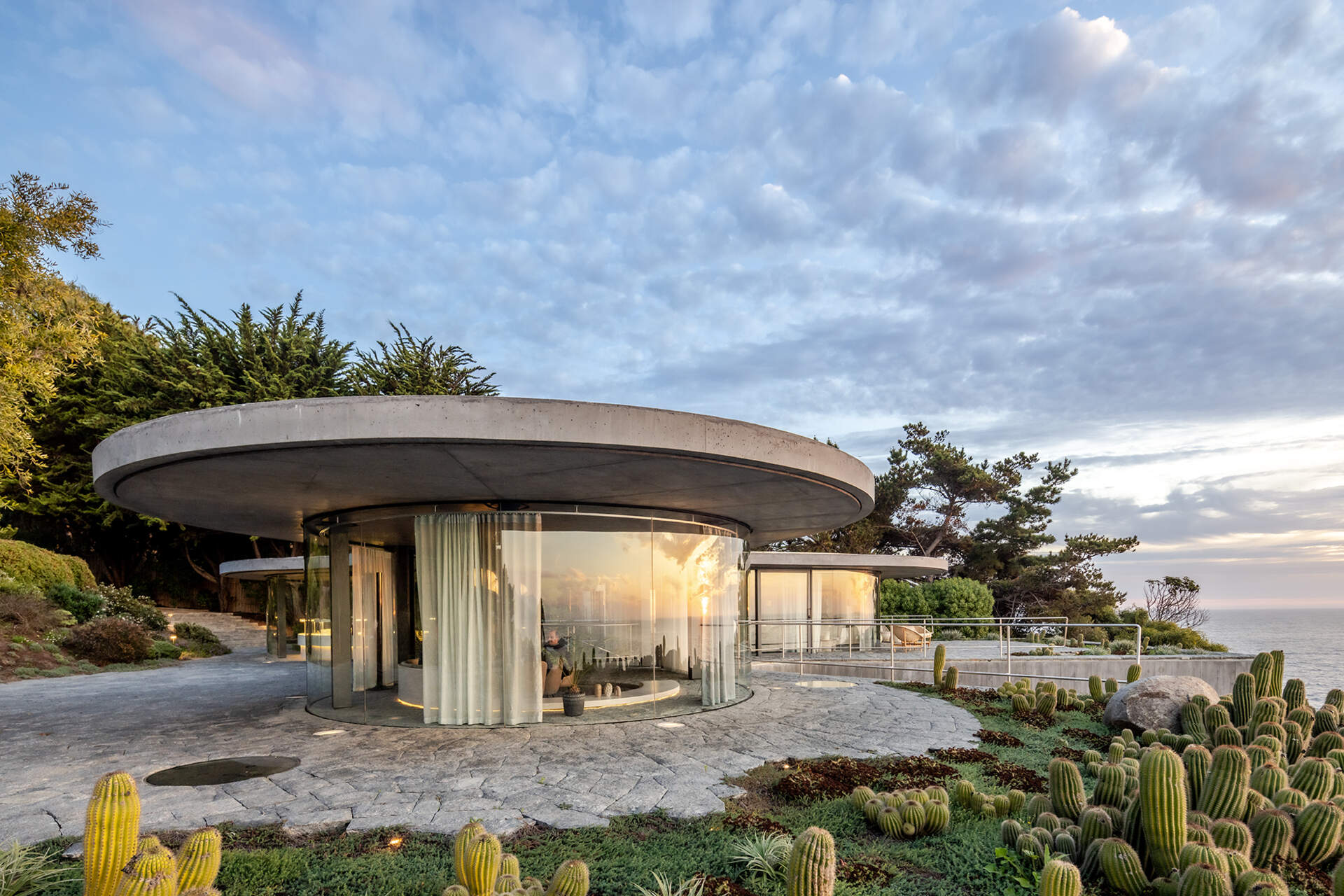 A concrete podium embedded into the landscape supports an amoeba-shaped glass pavilion, using stone, wood, and plank-form concrete throughout.