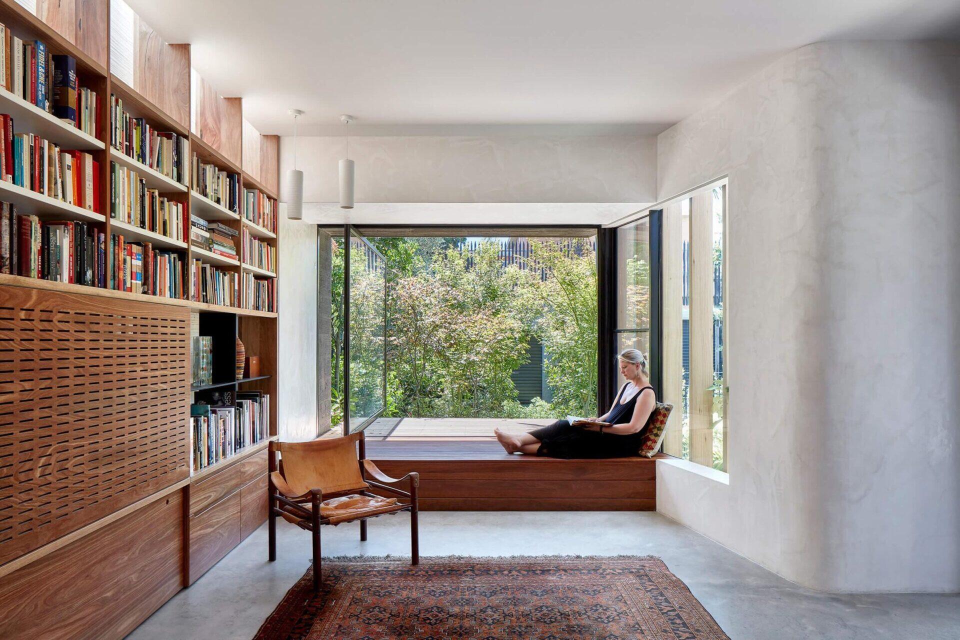 The living room centers on custom joinery. A wall of shelving wraps around the television, which is concealed behind a perforated panel to reduce visual clutter. A built-in window seat sits beside a large pivoting window, creating a focused place to sit and look out toward the rear yard.